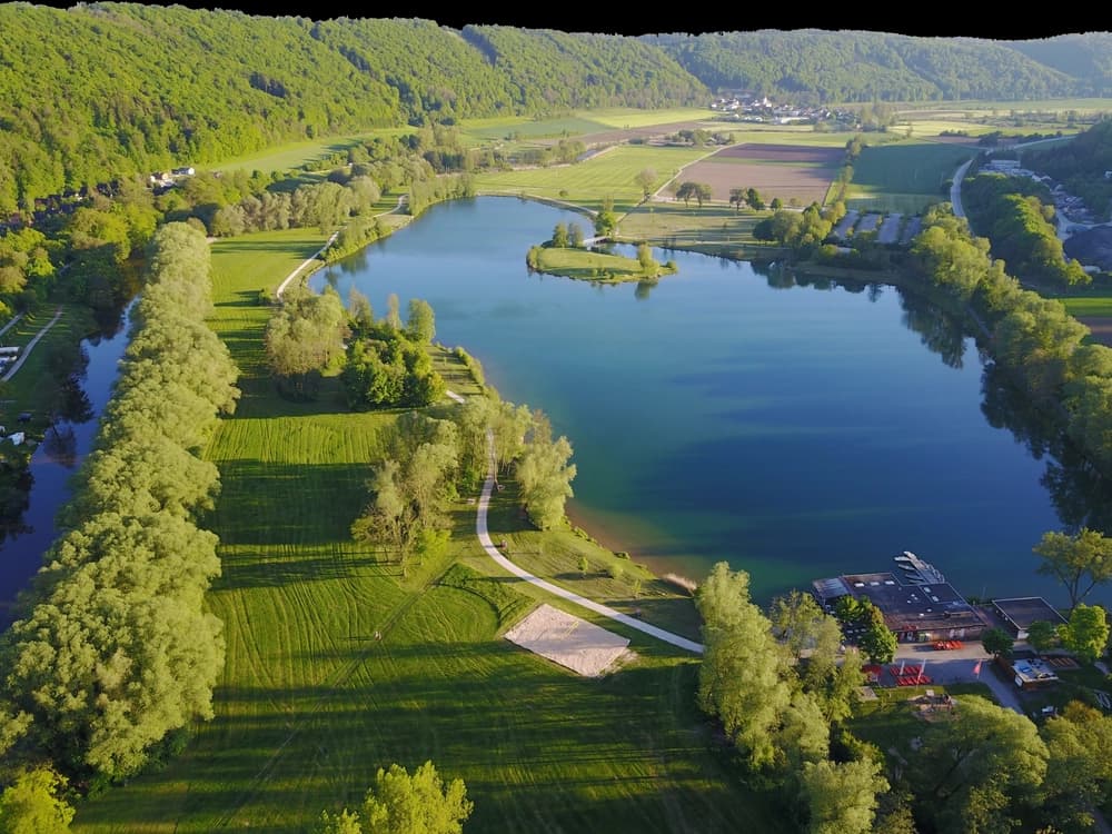 Luftaufnahme einer weitläufigen, sonnenbeschienenen Landschaft mit einem großen, tiefblauen See im Zentrum. Der See ist ringsum von üppigen grünen Wiesen und dichten Wäldern umgeben. Ein schmaler Kanal oder eine Ausbuchtung des Sees verläuft entlang der linken Bildseite, ebenfalls von Bäumen gesäumt. In der Mitte des Hauptsees befindet sich eine kleine, baumbestandene Insel. Entlang des unteren rechten Ufers stehen mehrere Gebäude mit dunklen Dächern, darunter ein Restaurant oder eine Freizeiteinrichtung mit Außenbestuhlung und roten Sonnenschirmen. Ein sandiger Volleyballplatz ist auf der Rasenfläche in Ufernähe zu sehen. Ein befestigter Weg schlängelt sich am Seeufer entlang und durch die Grasflächen. In der Ferne, hinter dem See und den Wäldern, erstrecken sich sanfte grüne Hügel und landwirtschaftliche Felder bis zu einer kleinen Häusergruppe, was auf eine ländliche Umgebung hindeutet. Der Gesamteindruck ist der einer friedlichen und malerischen Erholungsgegend.